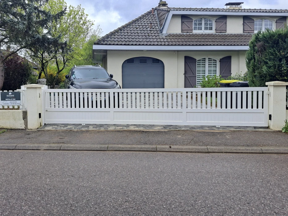 Portail coulissant blanc en PVC ou aluminium avec lames verticales ajourées, installé devant une maison pavillonnaire