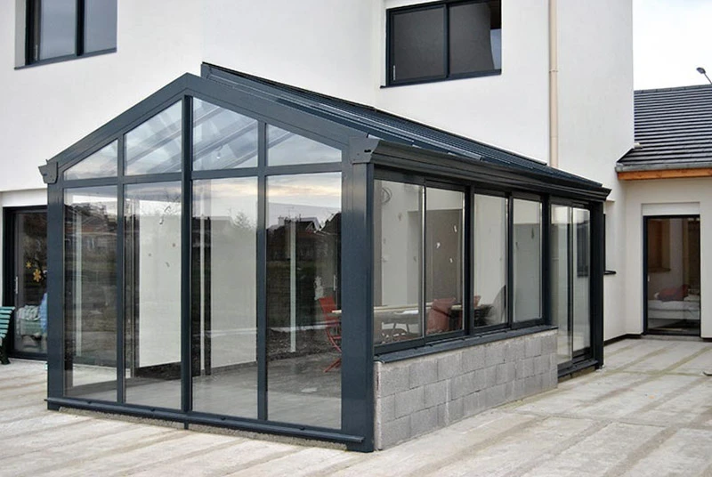 Salon contemporain dans véranda avec verrière, mobilier beige texturé disposé face à baies vitrées donnant sur paysage verdoyant