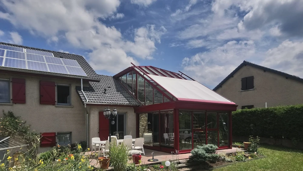 Salon contemporain dans véranda avec verrière, mobilier beige texturé disposé face à baies vitrées donnant sur paysage verdoyant