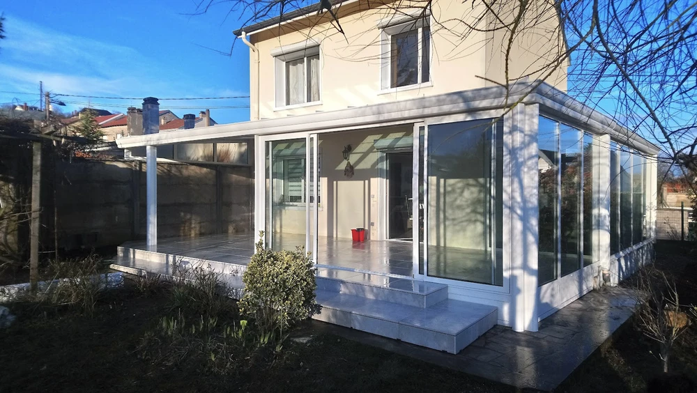 Salon contemporain dans véranda avec verrière, mobilier beige texturé disposé face à baies vitrées donnant sur paysage verdoyant