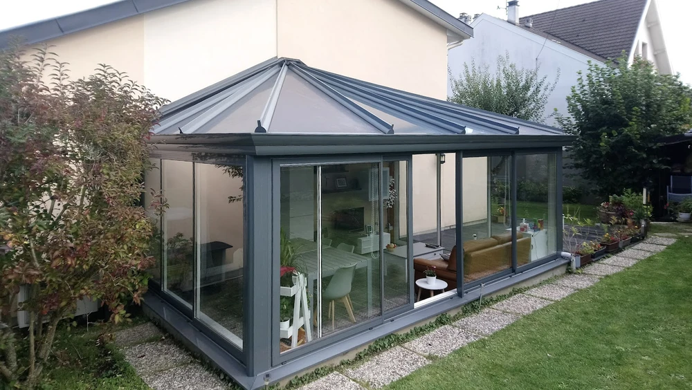 Salon contemporain dans véranda avec verrière, mobilier beige texturé disposé face à baies vitrées donnant sur paysage verdoyant