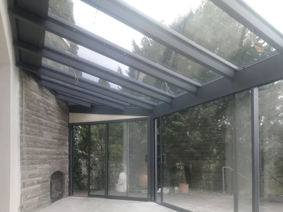 Salon contemporain dans véranda avec verrière, mobilier beige texturé disposé face à baies vitrées donnant sur paysage verdoyant